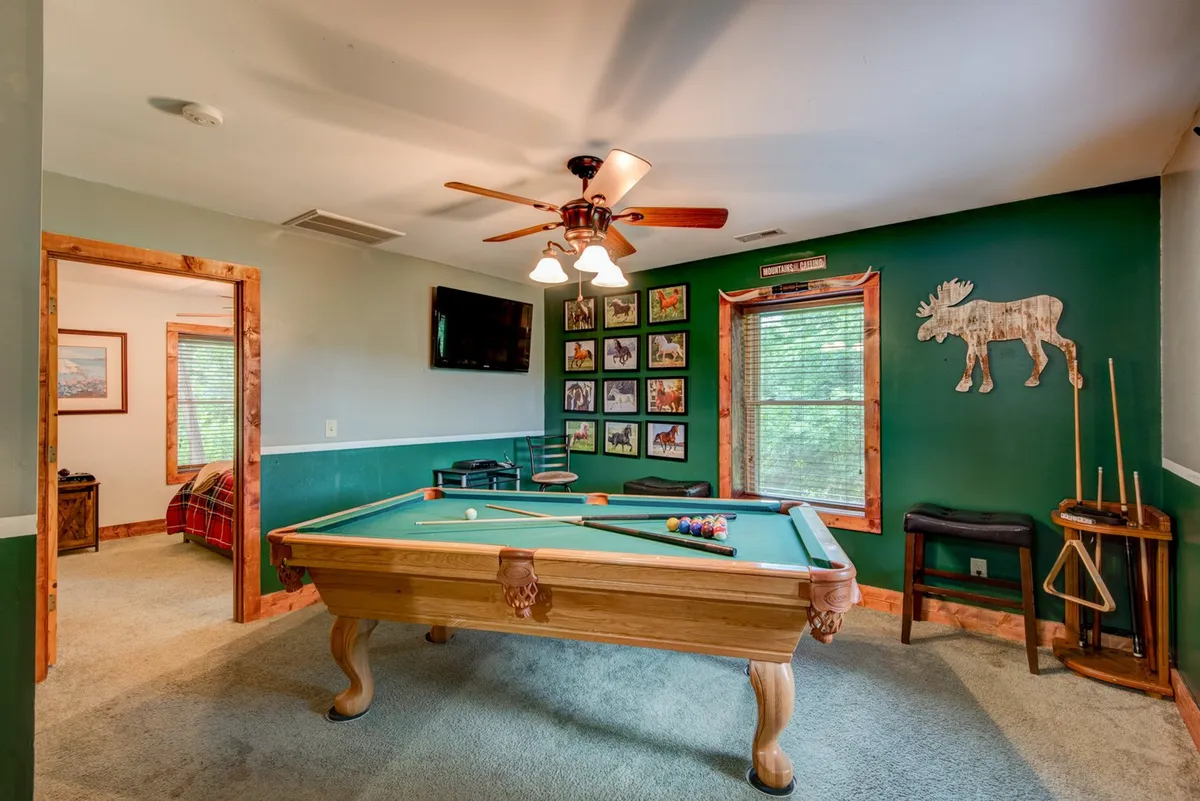 A spacious lower-level game room featuring a pool table at the center, vibrant wall decorations, a ceiling fan, and a television mounted on the wall. The arrangement is ideal for friendly pool tournaments or relaxed family game nights.