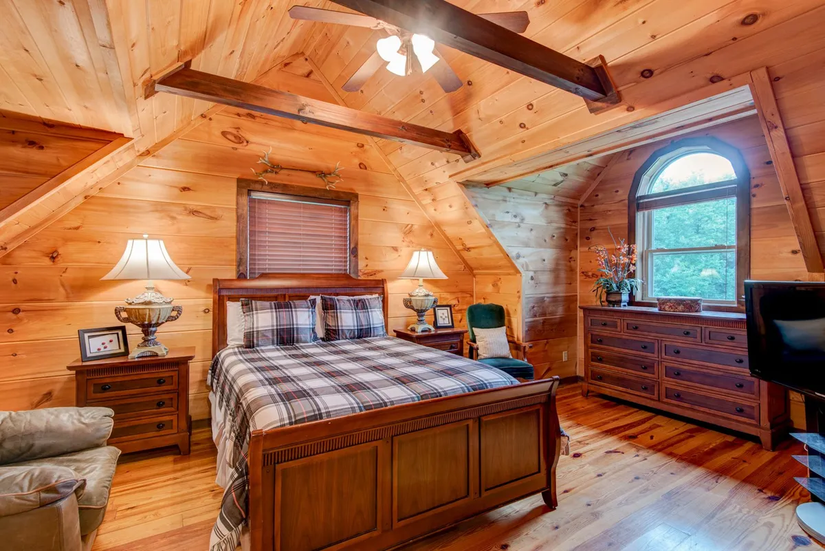 This image features a wooden bed frame with plaid bedding, flanked by table lamps and set beneath an arched window. The setting exudes warmth and rustic charm, with the window offering a view of nature and flooding the room with soft light.
