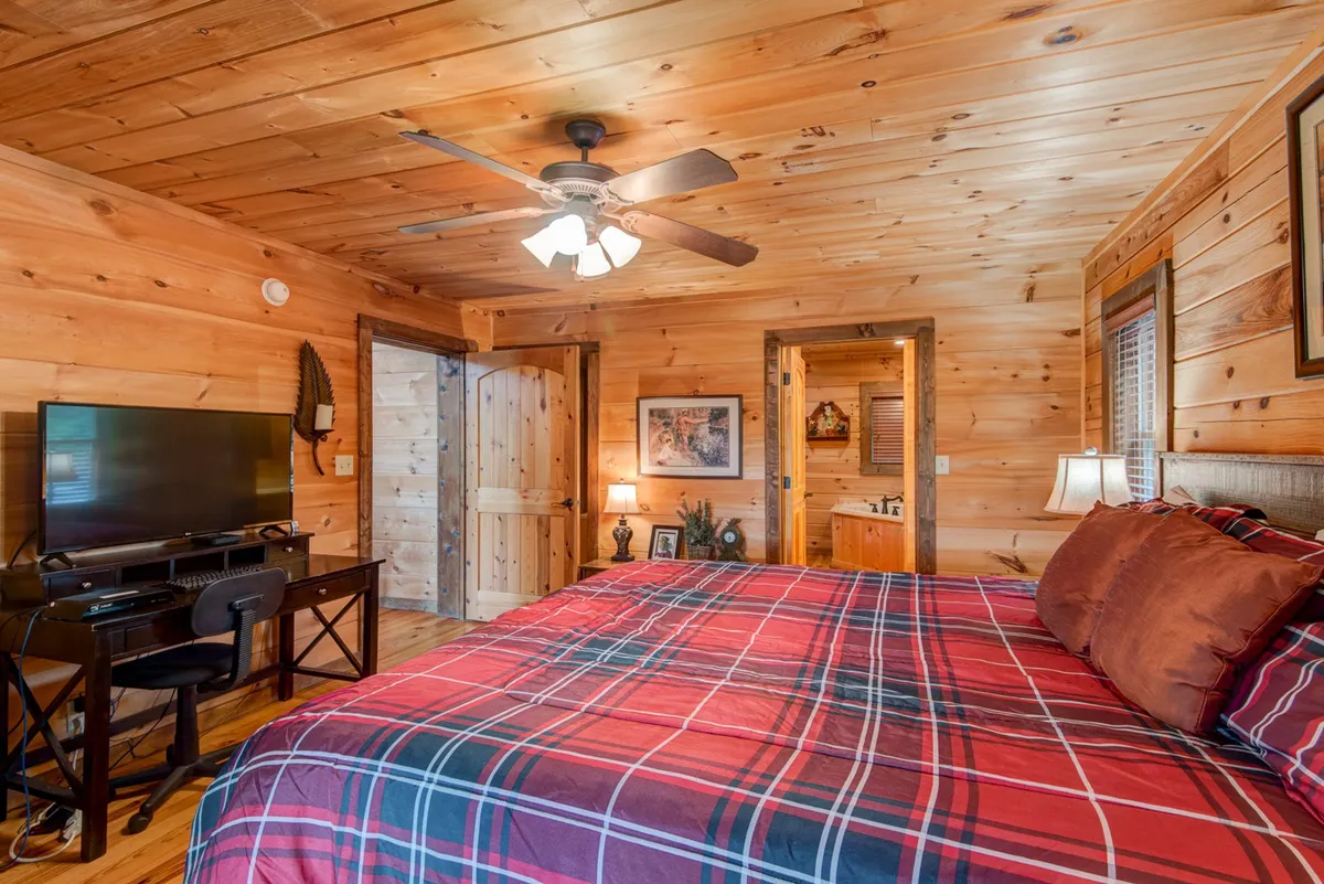 A queen bedroom with plaid bedding, wooden walls, ceiling fan, television, and a desk and chair. The space feels warm and inviting, ideal for rest or a quiet moment with a book.