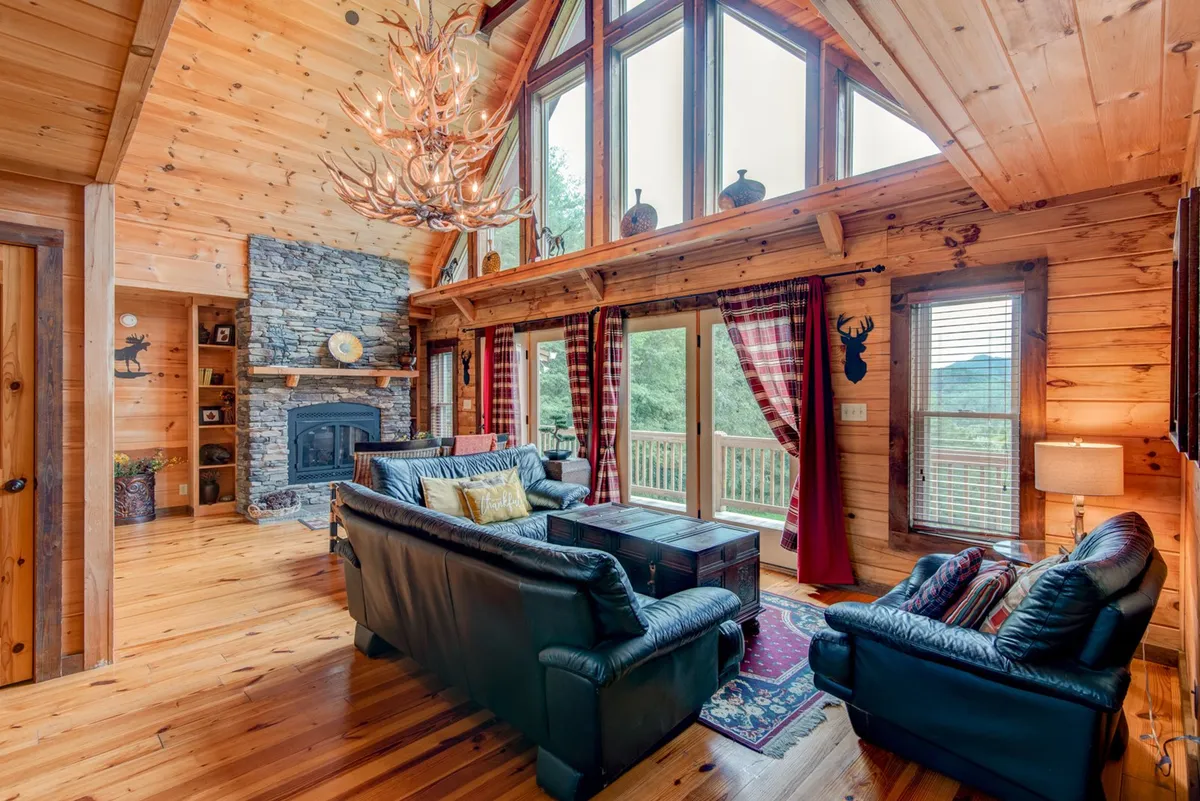 A spacious living room with soaring ceilings, expansive wall-to-wall windows showcasing mountain vistas, a majestic stone fireplace, a chandelier, and plush seating arranged for conversation or relaxation. Natural light floods the room, and the inviting arrangement encourages togetherness.