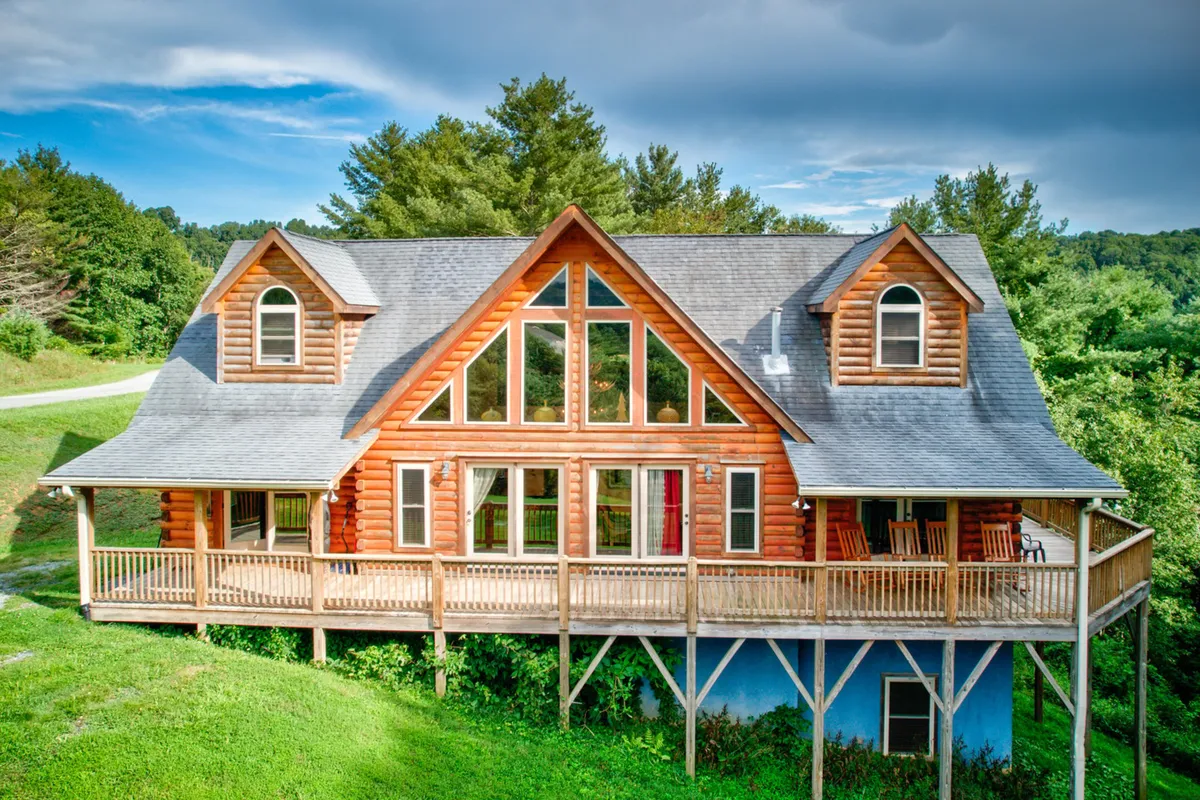 An exterior shot of the log cabin's expansive front porch, featuring large windows and surrounded by lush trees. The inviting porch encourages guests to relax outdoors and highlights opportunities for family gatherings, barbecues, and unwinding amid the scenic mountain landscape.