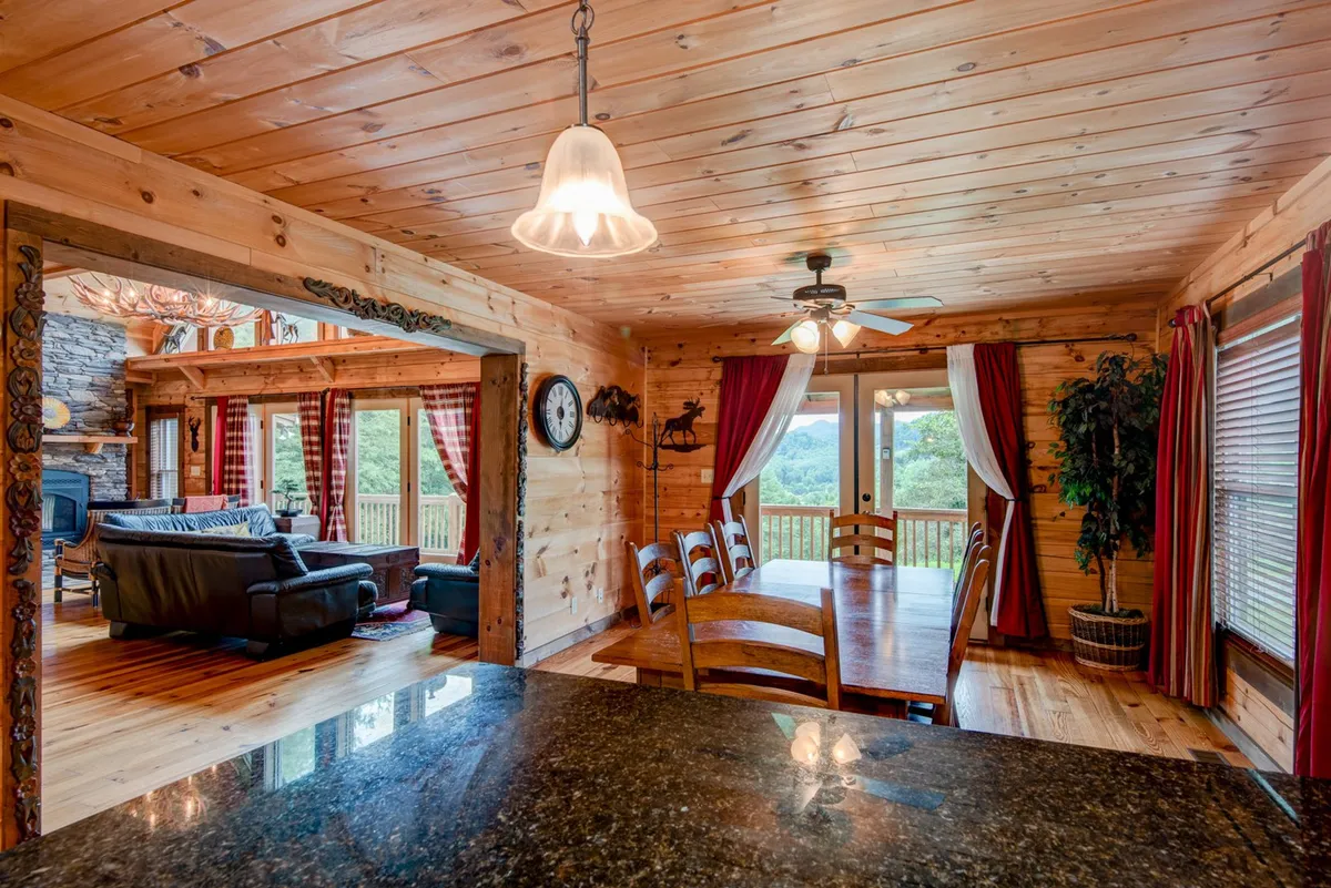 An open-concept area with a wooden dining table set for five, comfortable seating nearby, and large windows that let in abundant light and scenic views. Exposed wooden beams and ceiling add to the rustic ambiance, blending the dining and living areas seamlessly.