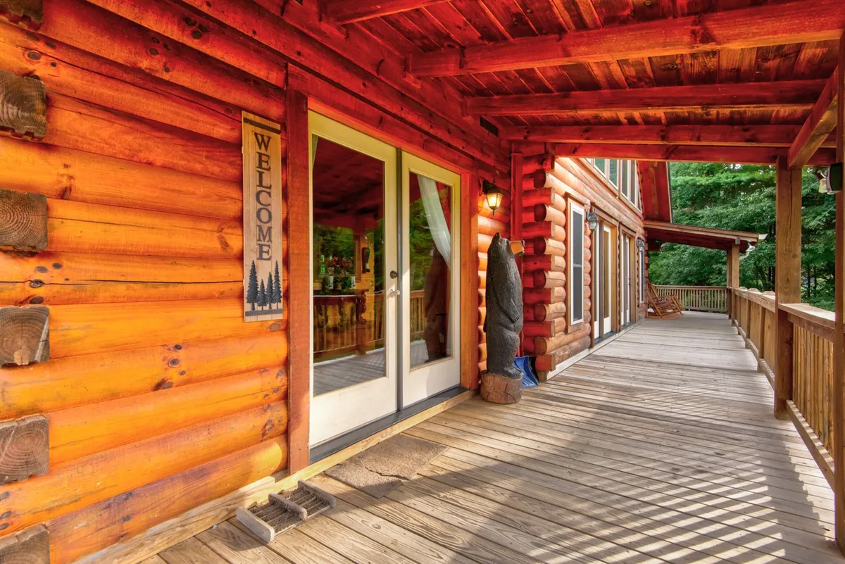 This exterior image showcases the log cabin structure with a welcoming sign, large glass doors, and a spacious wooden deck surrounded by nature. The setting conveys privacy and tranquility, while also hinting at the luxurious mountain experience awaiting inside.