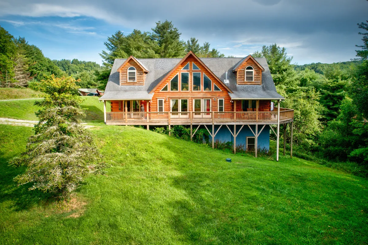 The image shows the exterior of a spectacular log cabin surrounded by lush greenery and towering trees. The home features expansive windows that let in natural light, a spacious wooden deck perfect for relaxation or gatherings, and a manicured lawn inviting outdoor activities. The tranquil setting is enhanced by the serene mountain atmosphere, making it an ideal retreat for families or friends.