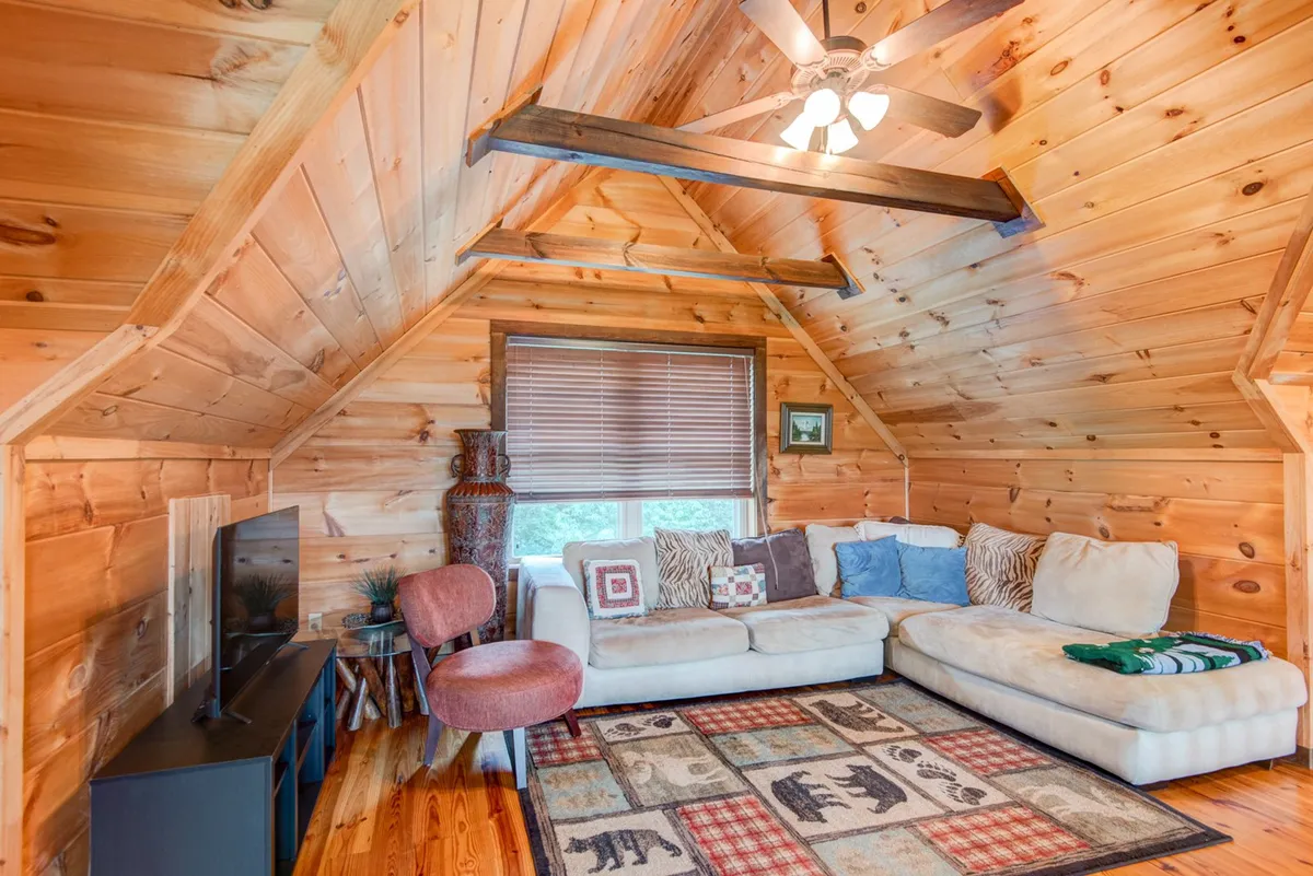 A cozy loft space with a comfortable sofa, patterned rug, wooden walls, ceiling fan, and a window letting in soft light. The room invites relaxation, reading, or watching TV, aligning with quieter moments away from the action.