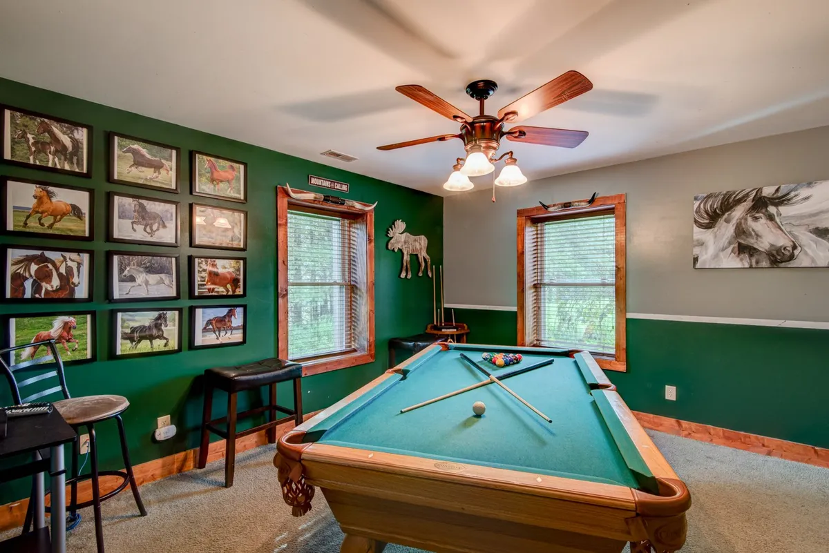 A welcoming game room with a pool table beneath bright lighting, horse-themed artwork on the wall, and several windows that bring in natural light. The setting feels open, social, and ready for friendly competition or casual relaxation.