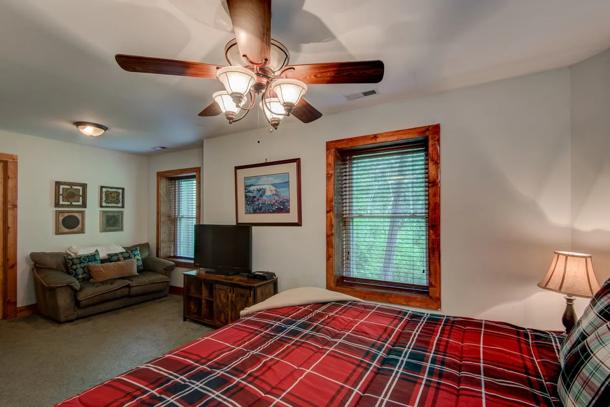 A cozy upstairs sitting room features a plaid bedspread, ceiling fan, sofa, and artwork on the walls. The space is inviting and multifunctional, ideal for relaxing, watching television, or sharing stories with loved ones.