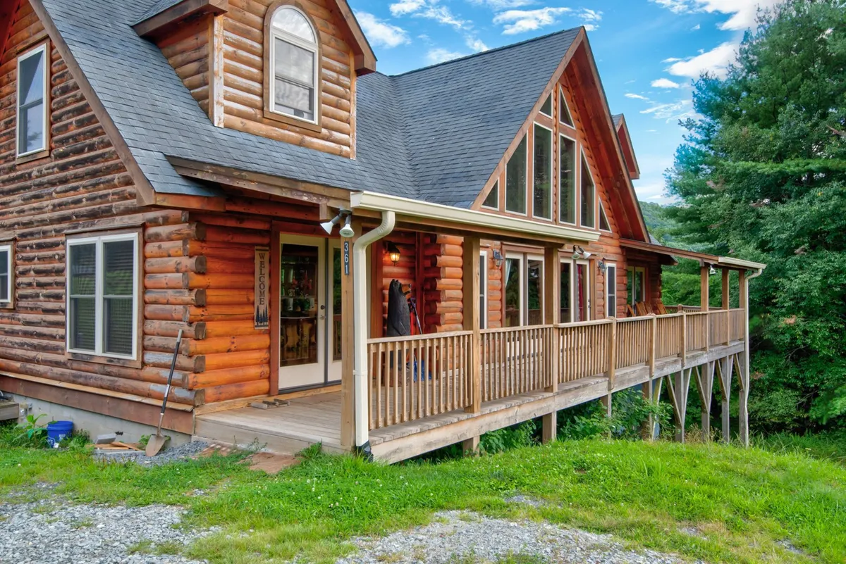 A welcoming log cabin structure with a large front porch and gabled roof, surrounded by lush greenery. The porch offers a cozy spot for outdoor gatherings, with ample space for seating and a stunning view of the mountains, especially at sunset or during the early morning.